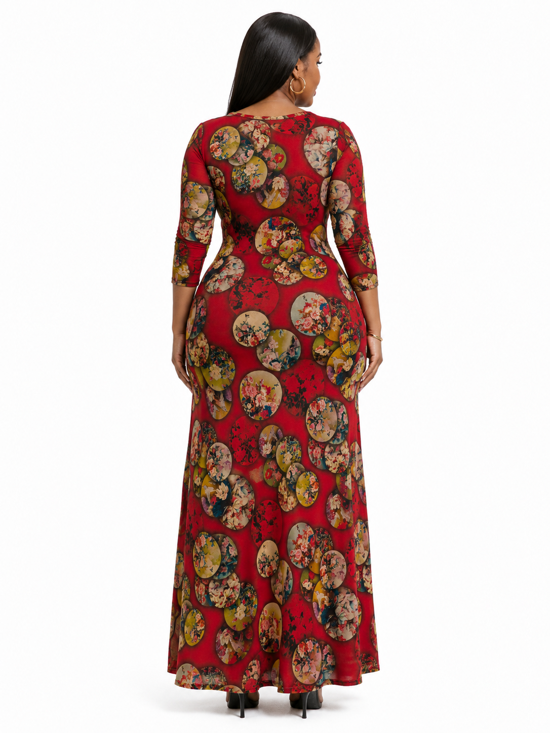 A woman viewed from the back wearing a floor-length red dress with three-quarter sleeves and an allover circular floral print in muted yellow, green, and beige tones, photographed against a plain white studio background.
